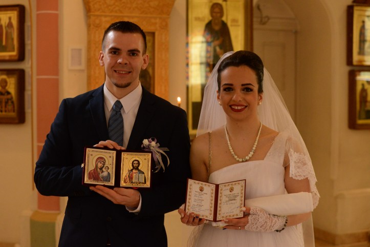 Us the married couple holding an icon and the wedding certificate by the church