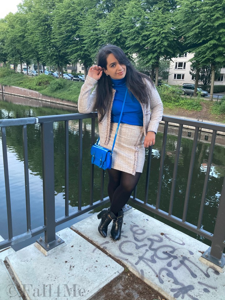 A beige skirt suit in French tweed worn with black accessories and an azure blue turtleneck and bag.