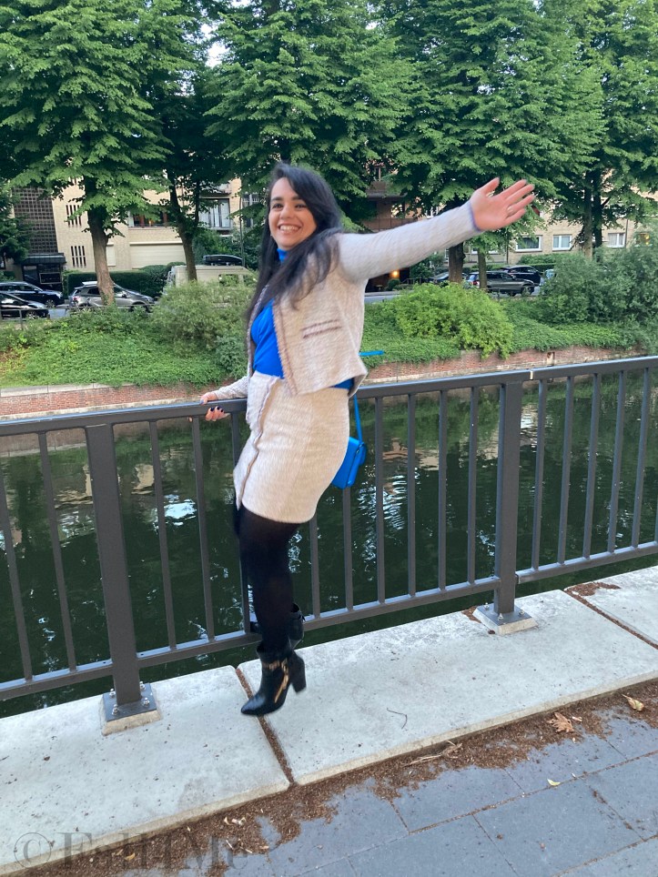 A beige skirt suit in French tweed worn with black accessories and an azure blue turtleneck and bag.