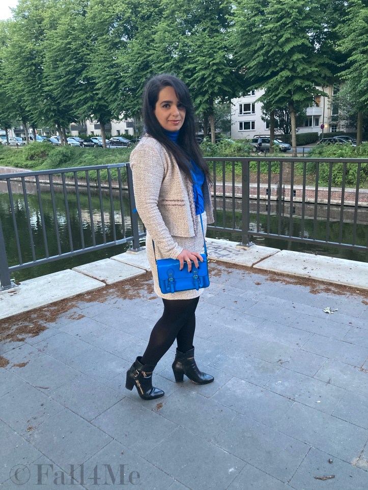 A beige skirt suit in French tweed worn with black accessories and an azure blue turtleneck and bag.
