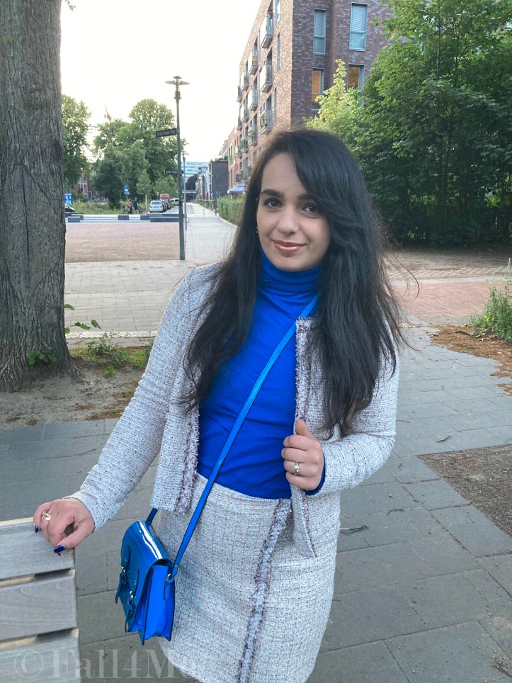 A beige skirt suit in French tweed worn with black accessories and an azure blue turtleneck and bag.