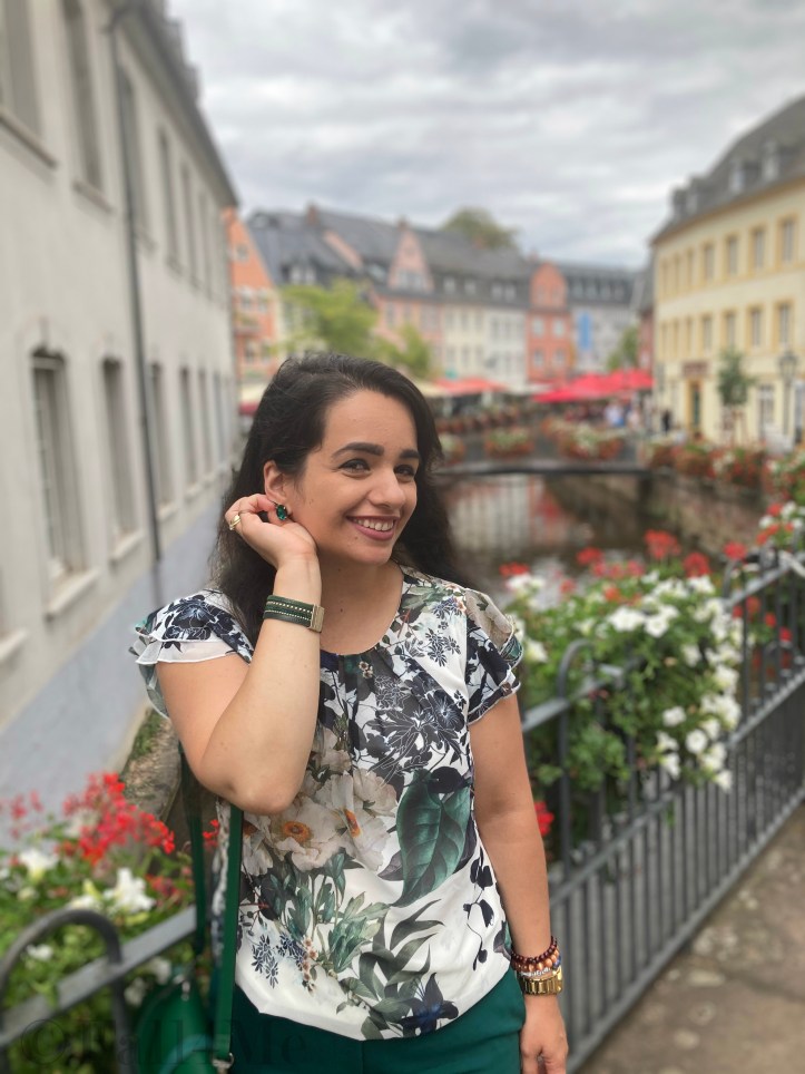 Green business pants combined with a white blouse with roses and leafs on it, a green cross body bag and golden accessories make the perfect urban jungle look.